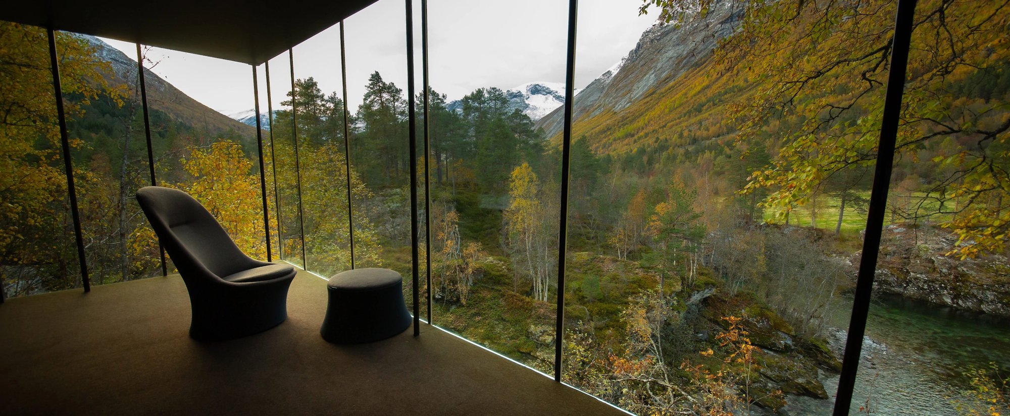 A lounge chair facing a floor-to-ceiling glass wall that looks onto the river and autumn forest. Photo: Juvet Landscape Hotel