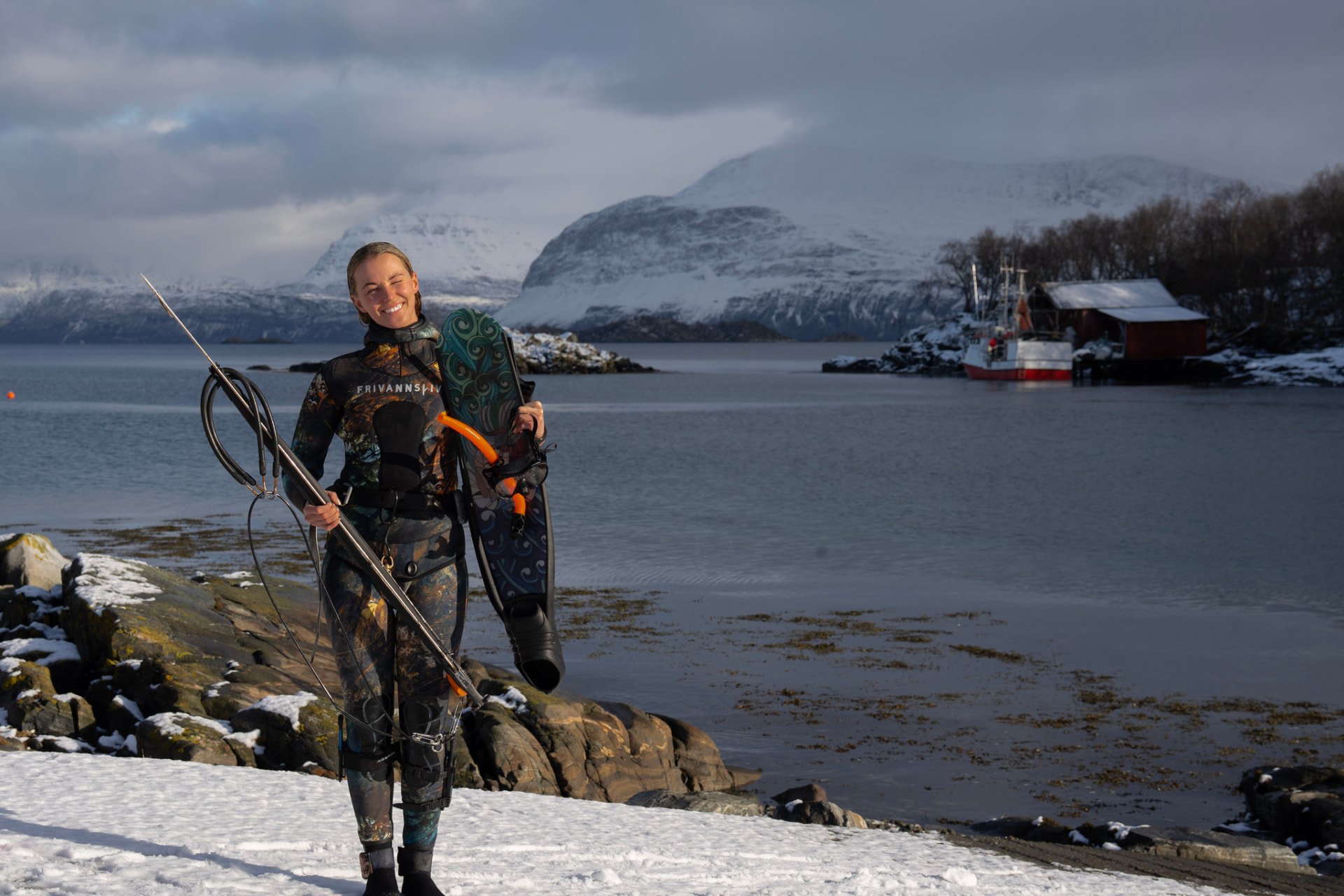 A smiling spearfisher with fins and speargun on a snowy Norwegian shore. Photo: Frivannsliv