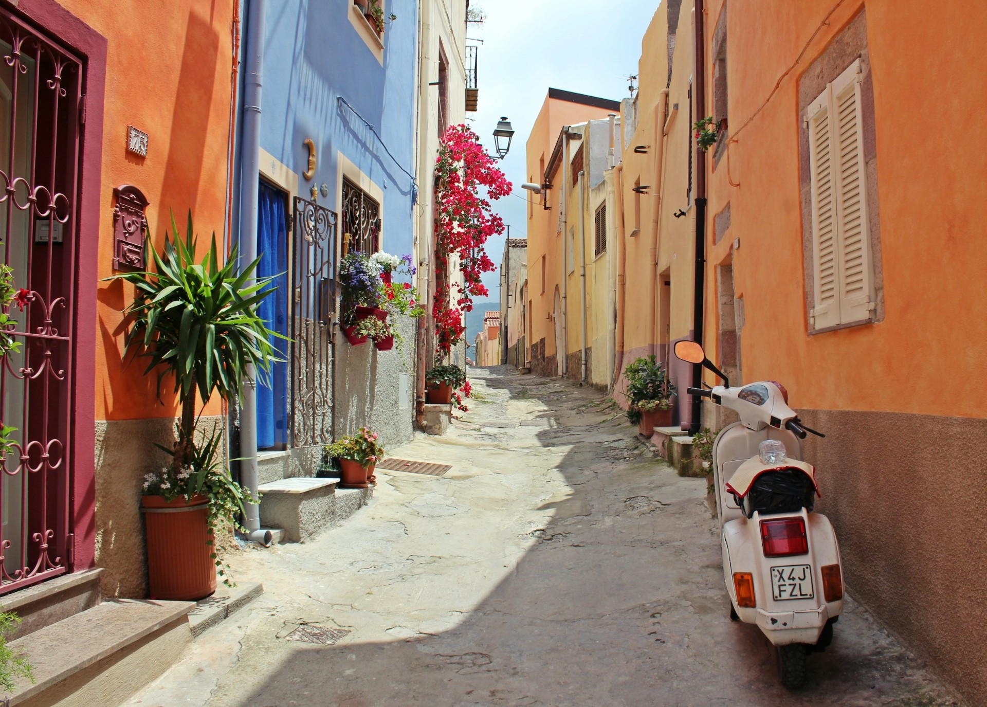 A white Vespa parked in a bougainvillea-hung Sardinian alley of orange, blue and yellow houses